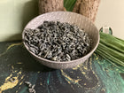 A ceramic bowl filled with dried, curled dark gray-green herbal tea leaves, set on a dark green and gold marbled surface with decorative green leaves and tree trunks in the background.