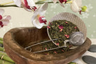 Tea leaves in a wooden bowl with a metal scoop and orchids in the background