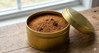Gold cylindrical container with a lid on a wooden surface, filled with a brown powder.